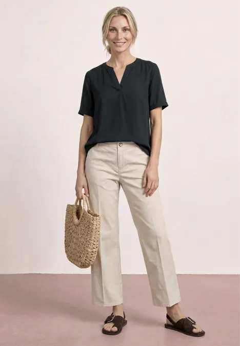 Frau in schwarzer Bluse, beiger Hose, schwarzen Sandalen und Korbtasche vor schlichtem Hintergrund.