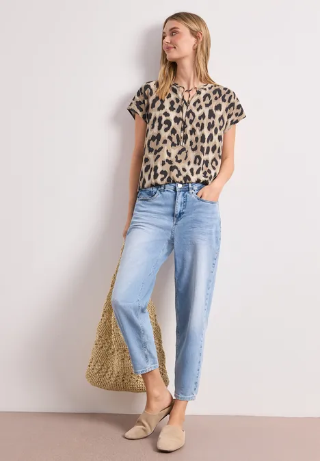 Frau vor weißem Hintergrund trägt Leopardenbluse, hellblaue Jeans, Basttasche und Espadrilles.