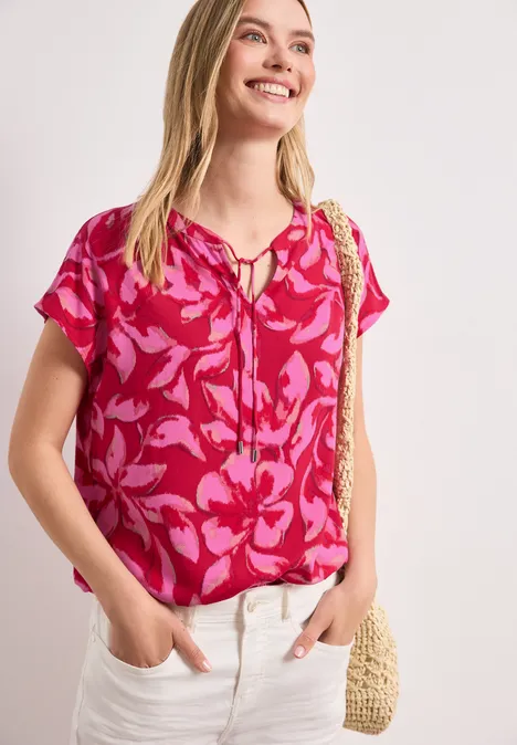 Eine Frau in pinker Bluse mit Blumenmuster, heller Hose und Strohtasche.