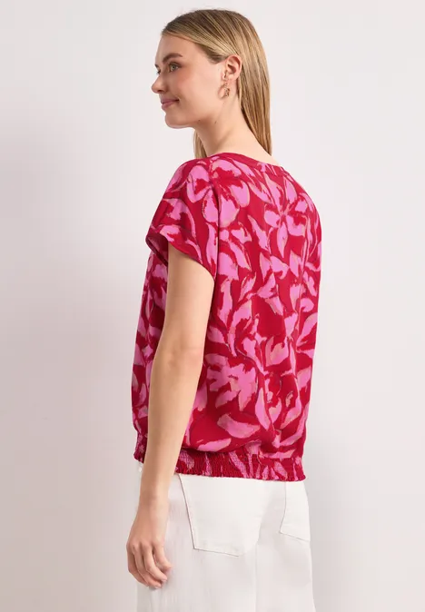 Frau von der Seite in rosa Bluse mit Blättermuster und weißen Hosen.