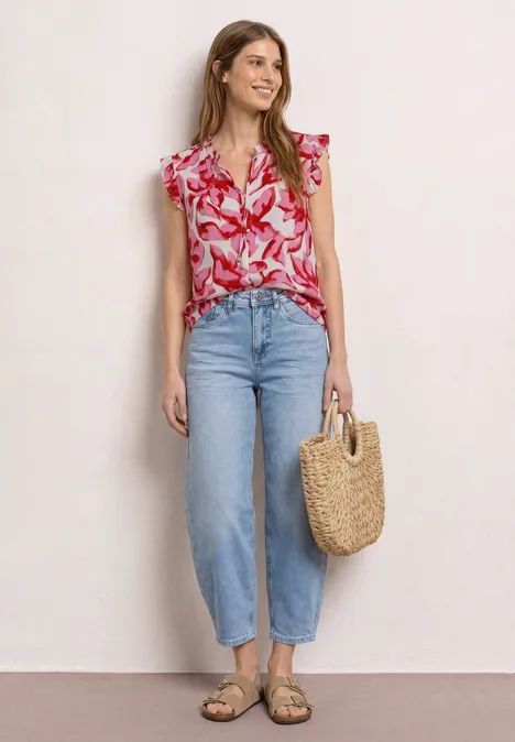 Vrouw in rood gebloesd shirt, lichtblauwe high-waist jeans, sandalen en rieten tas voor lichte muur.