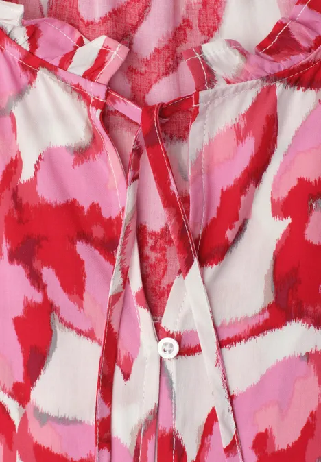 Close-up van een blouse met abstract rood, roze en wit patroon, V-hals en knoopdetail.