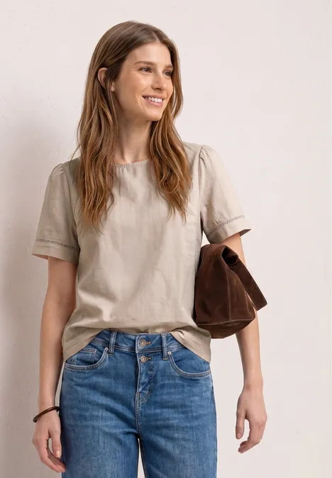 Een persoon in een beige T-shirt en spijkerbroek, met een bruine tas over de arm, tegen een neutrale achtergrond.