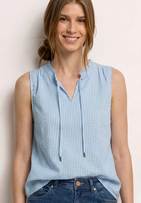 Vrouw met mouwloos lichtblauw-wit gestreept topje voor een neutrale achtergrond.
