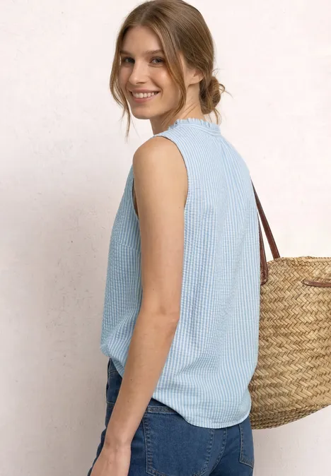 Zijaanzicht van een vrouw met lichtblauwe mouwloze top en jeans, met een grote gevlochten strooien tas op de schouder.