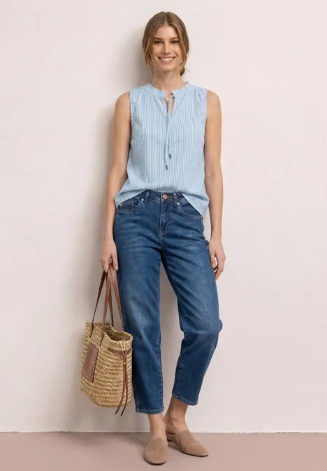 Vrouw in lichtblauwe mouwloze top, blauwe jeans en rieten tas tegen neutrale achtergrond.