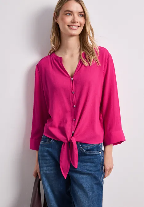 Vrouw in roze blouse geknoopt in de taille en blauwe jeans voor een lichte achtergrond.