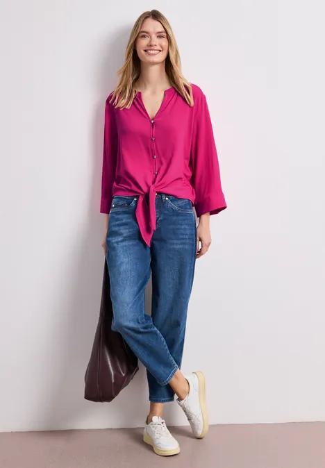 Vrouw in fuchsia blouse met knoop voor, losse blauwe jeans en witte sneakers tegen een lichte muur.
