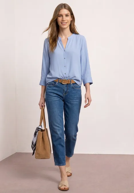 Frau in hellblauer Bluse, dunkelblauen Jeans, braunem Gürtel, Sandalen und Handtasche.