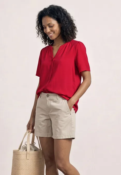 Eine Person trägt ein rotes Kurzarmshirt, beige Shorts und hält eine Strohtasche.