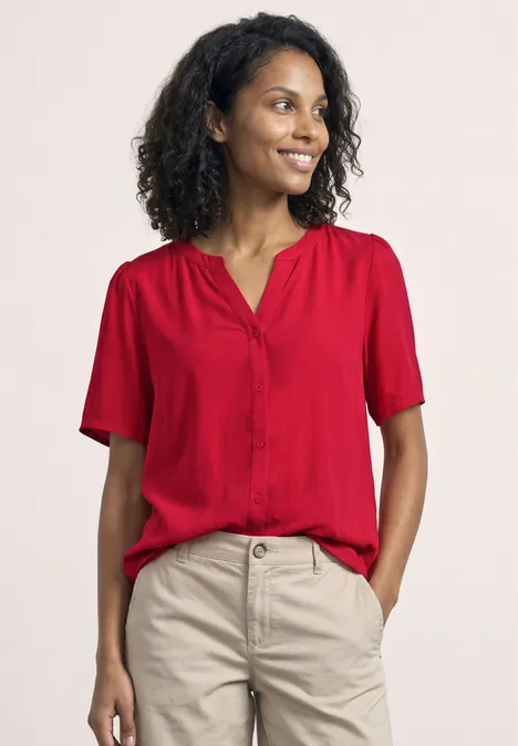 Eine Frau mit lockigem Haar trägt eine rote Bluse und helle Hose.