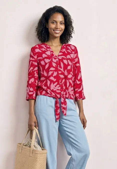 Vrouw draagt rode gebloemde blouse met knoop, lichtblauwe broek en beige tas.