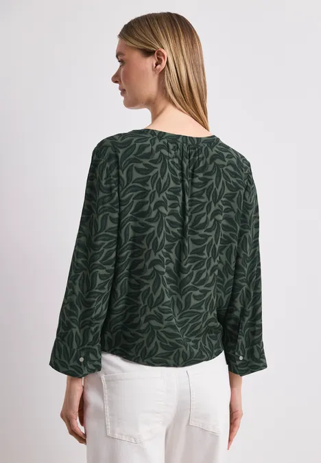 Vrouw van achteren, gekleed in een donkergroene blouse met bladpatroon en een witte broek.