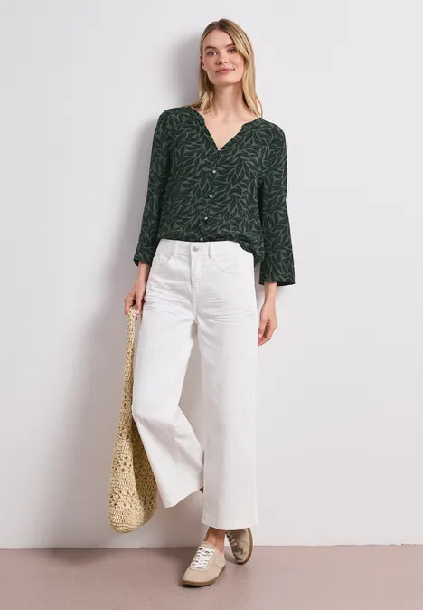 Vrouw in donkergroene blouse, witte wijde broek, gouden schoenen en rieten tas voor lichte achtergrond.