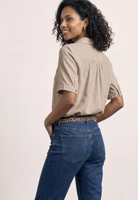 Chemisier à manches courtes en couleur unie clay sand Frau mit lockigem Haar trägt beigefarbenes Hemd und blaue Jeans, steht mit roten Nägeln in den Taschen, Rückenansicht.