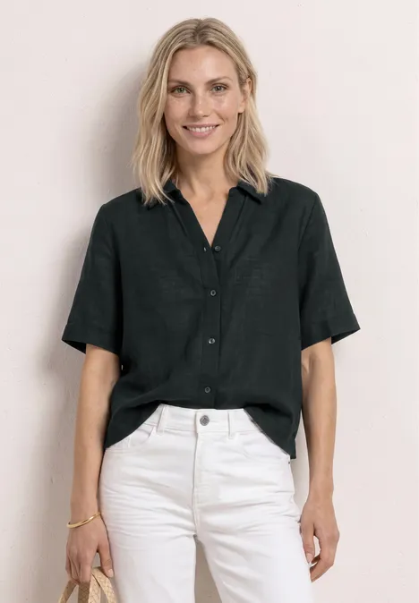 Vrouw met blond haar draagt zwarte korte mouw blouse en witte jeans voor een lichte achtergrond.
