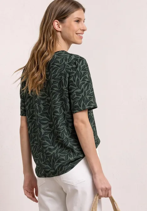 Vrouw van achteren in groen t-shirt met bladprint en witte broek tegen neutrale achtergrond.