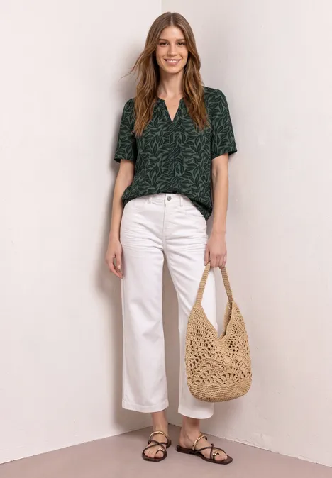 Vrouw in groene top, witte broek, zwarte sandalen en beige gehaakte tas.