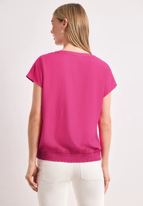 Frau, Rückansicht, pinkes Kurzarmshirt mit Gummizug am Saum, weiße Hose, weißer Hintergrund.
