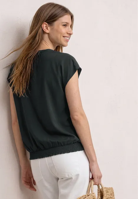 Frau mit schulterlangem Haar, schwarzem Shirt, weißer Hose und Strohtasche steht vor heller Wand.