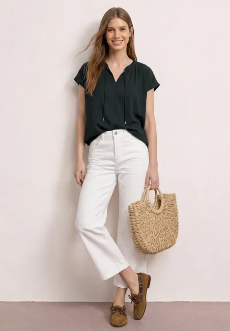 Frau in schwarzem Oberteil und weißen Jeans, mit braunen Loafers und Strohtasche vor hellem Hintergrund.