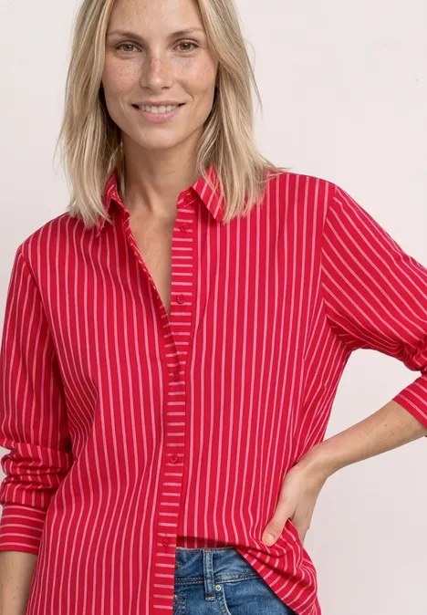 Blonde vrouw draagt rood overhemd met witte strepen, licht glimlachend, hand in de heup.