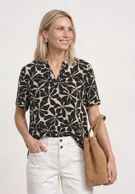 Frau in schwarzer Bluse mit weißem Blumenmuster, weißer Hose und beiger Tasche vor weißem Hintergrund.