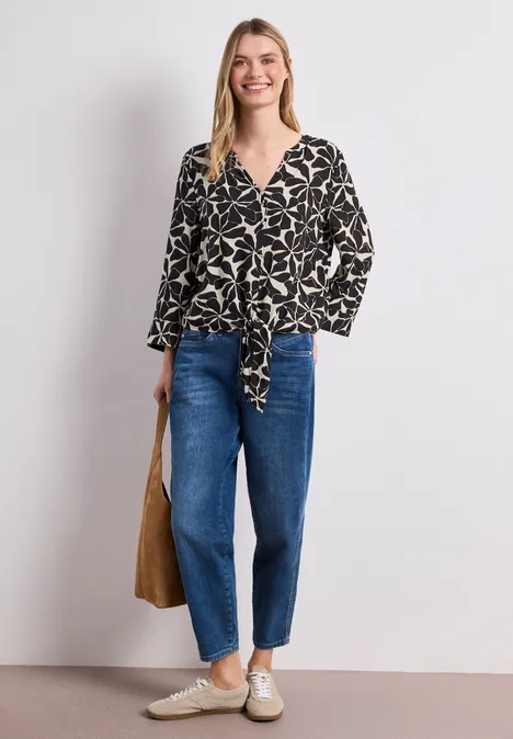 Vrouw draagt zwart bloesje met wit dessin, blauwe jeans, lichte sneakers en een bruine tas.