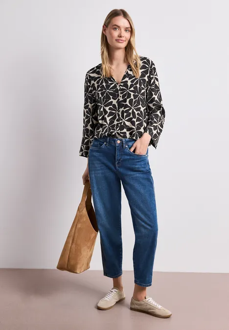 Vrouw met motiefblouse, blauwe jeans en beige schoenen houdt grote bruine tas voor een witte achtergrond.