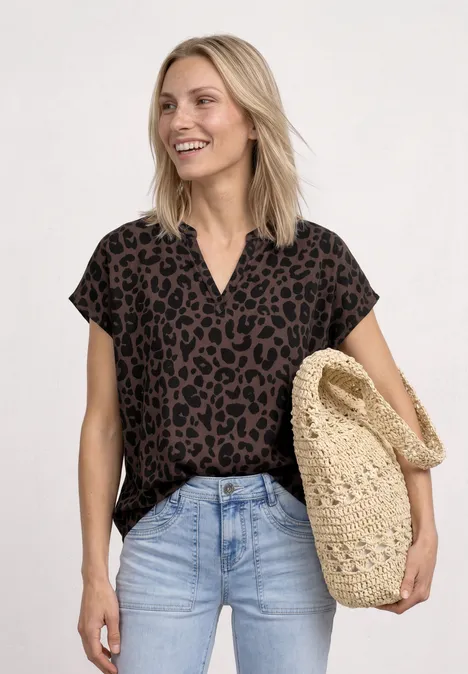 Vrouw met bruin luipaardprintblouse, lichtblauwe jeans en een gevlochten strozak voor een lichte achtergrond.