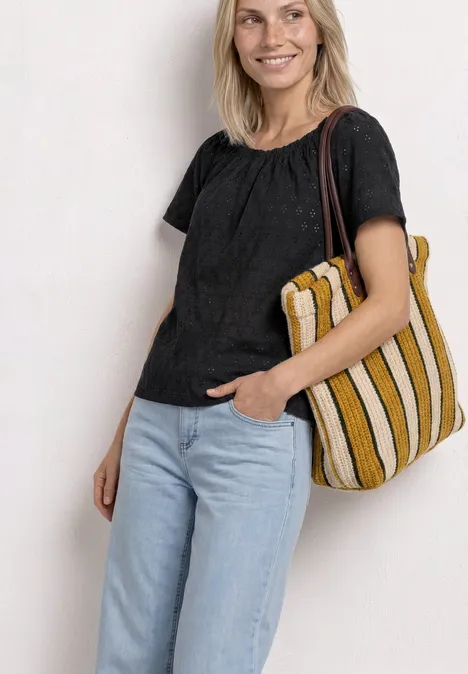 Frau trägt schwarze Bluse, hellblaue Jeans und hält eine gelb-weiße gestreifte Tasche lässig über der Schulter.