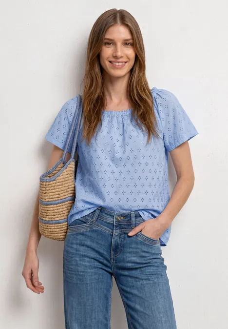 Carmen blouse cashmere blue Frau trägt hellblaues, kurzärmeliges Oberteil mit Muster, Jeans und hält eine geflochtene Korbtasche.