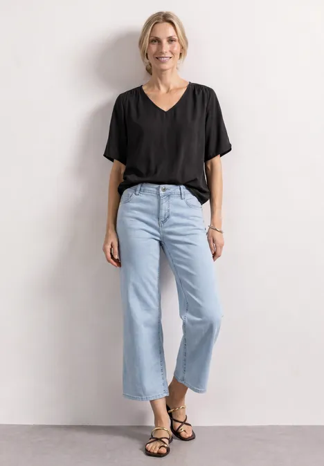 Person in lockerem schwarzen T-Shirt, hellblauen weiten Jeans und schwarzen Sandalen vor weißem Hintergrund.