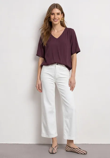Femme en T-shirt violet col V, jean blanc raccourci et ballerines zébrées devant un mur neutre.