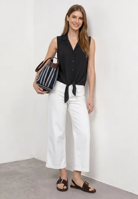 Blusentop mit Bowling-Kragen und Knoten Black Frau trägt schwarzes ärmelloses Top mit Knotendetail, weiße Culottes, schwarze Sandalen und gestreifte Tasche.