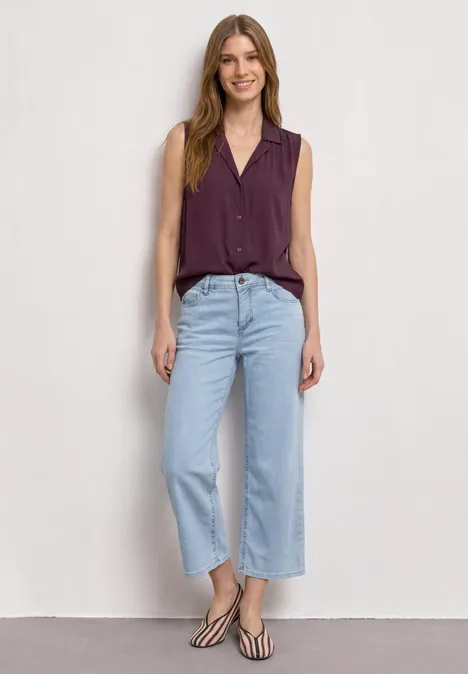 Blusentop mit Bowling-Kragen und Knoten rustic rouge Frau in ärmelloser lila Bluse, hellblauen Culotte-Jeans und Sandalen vor weißem Hintergrund.