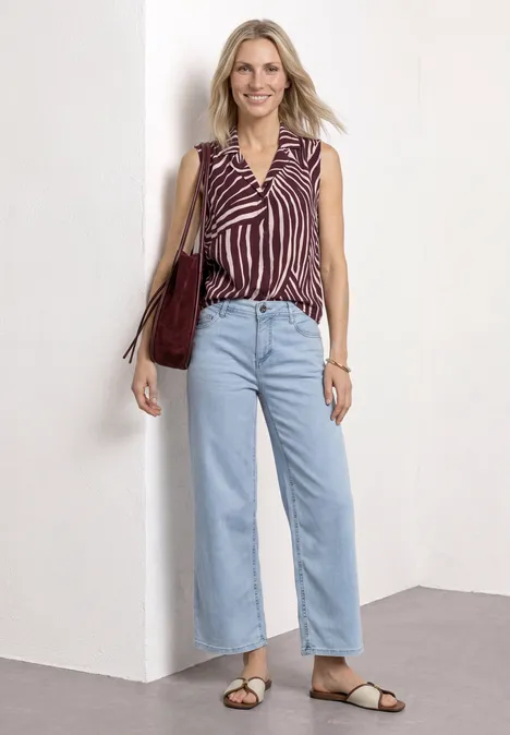 Blusentop mit Bowling-Kragen und Knoten rustic rouge Frau steht vor weißer Wand, trägt gestreifte ärmellose Bluse, helle Jeans, Sandalen und eine rote Schultertasche.