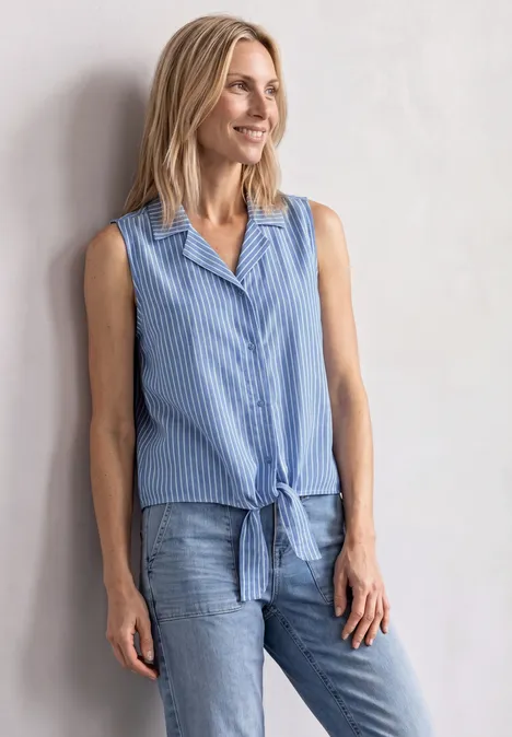 Vrouw in mouwloos, blauw-wit gestreept hemdje met knoop voor en spijkerbroek tegen lichte muur.