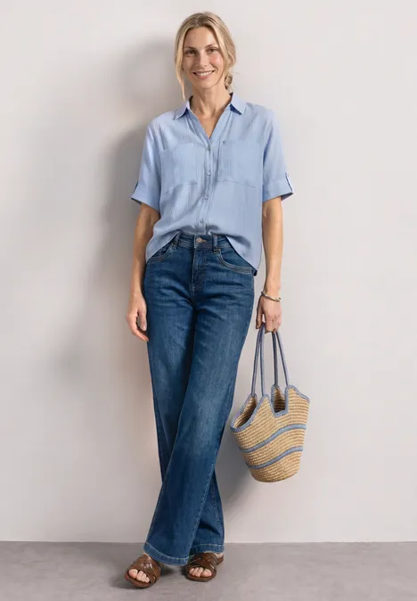 Kurzarm Hemdbluse mit Turn-Up-Details cashmere blue Frau in hellblauer Bluse, weiten Jeans und Sandalen hält eine gestreifte Strandtasche vor neutralem Hintergrund.