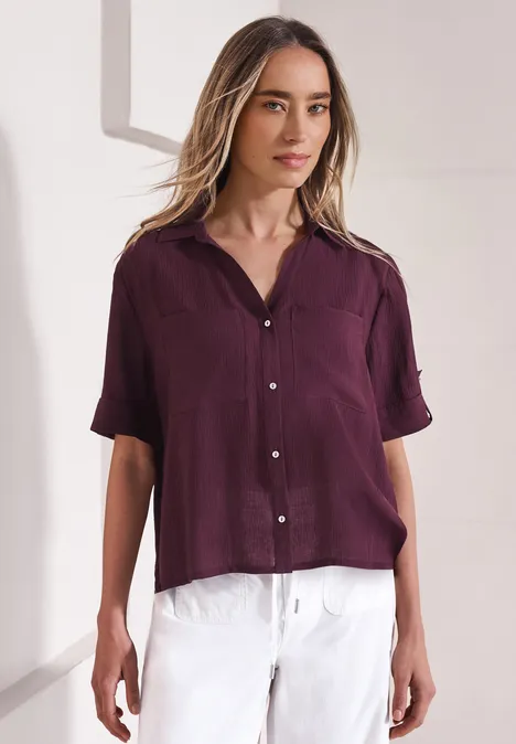 Vrouw in een losse paarse blouse met korte mouwen en een witte broek tegen een lichte achtergrond.