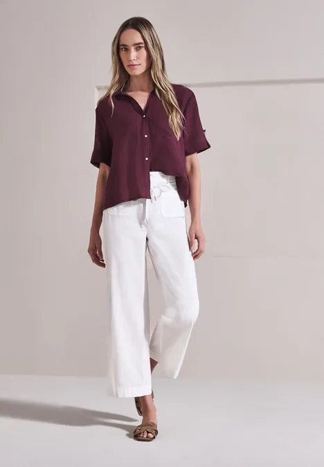 Vrouw tegen lichte achtergrond, bordeaux blouse, witte wijde broek en sandalen.
