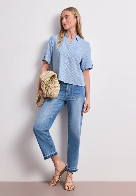 Kurzarm Hemdbluse mit Turn-Up-Details original blue Frau in hellblauer Bluse, Jeans und Sandalen hält Strohhut in lässiger Pose vor hellem Hintergrund.