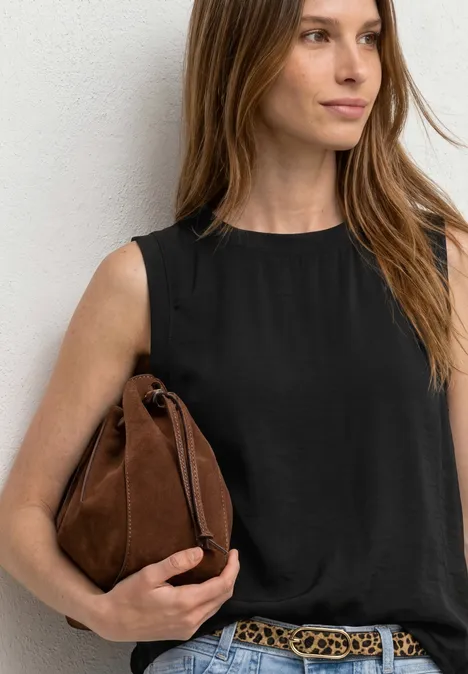 Schlichtes Outfit: Frau in schwarzem Top, brauner Tasche und Gürtel lehnt an weißer Wand.