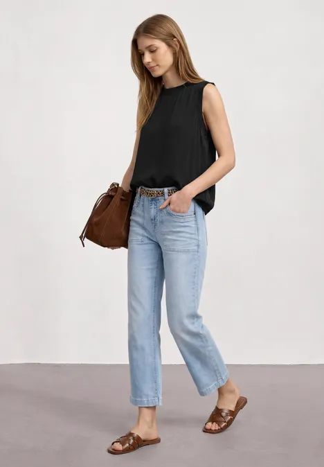 Frau in schwarzem Top, blauen Jeans und braunen Sandalen mit passender Tasche vor hellem Hintergrund.