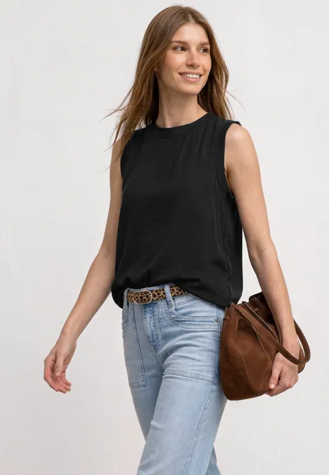 Frau trägt schwarzes ärmelloses Top, helle Jeans mit Gürtel und eine braune Umhängetasche vor weißem Hintergrund.