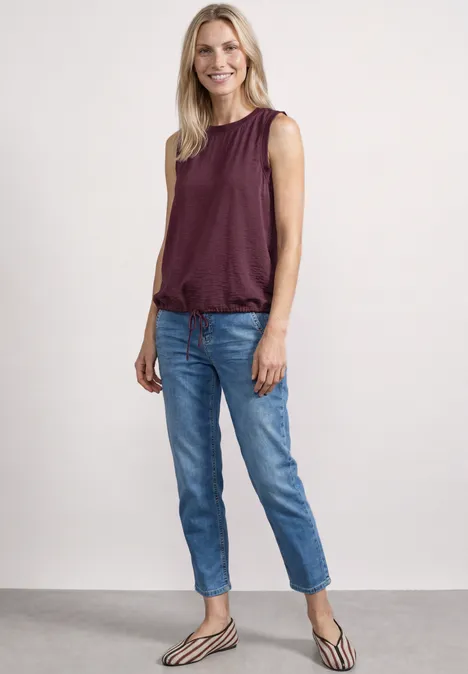 Blusentop mit Rundhals und Tape-Detail rustic rouge Eine Frau in ärmellosem bordeauxrotem Top, blauen Jeans und gestreiften Ballerinas steht vor hellem Hintergrund.