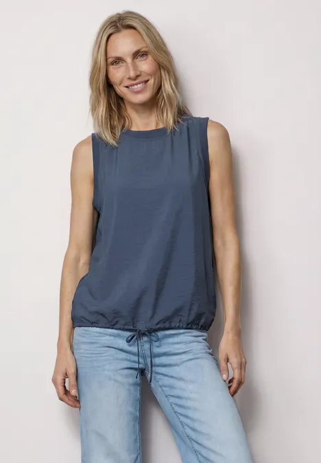 Frau mit ärmelloser, dunkelblauer Bluse und hellen Jeans steht vor hellem Hintergrund.