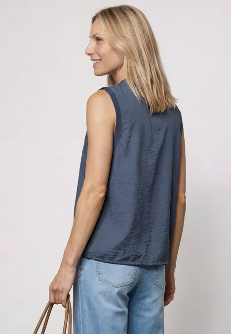 Frau mit blondem Haar von hinten, trägt ein ärmelloses dunkelblaues Oberteil, Jeans und hält eine kleine Tasche.