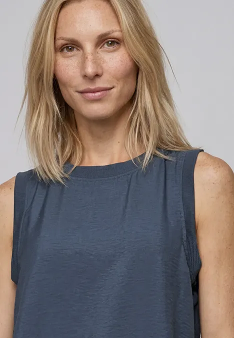Blonde Frau im ärmellosen, dunklen Oberteil vor hellem Hintergrund.