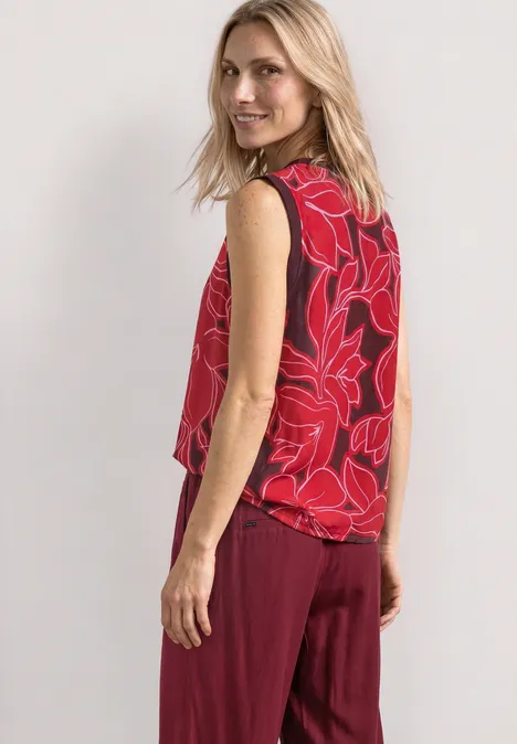 Frau in rotem ärmellosem Top mit Blumenmuster und weinroter Hose vor hellem Hintergrund.